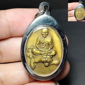 以下が含まれることがあります： 虎の上に座る僧侶の金色のレリーフが施された、涙型のペンダント。ペンダントは磨かれた銀色の金属フレームに収められており、上部にはチェーンやコードに取り付けるための円形のバチカンが付いています。
