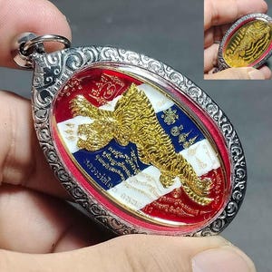 Może przedstawiać: Owalny srebrny wisiorek ze złotym wzorem tygrysa. Wisiorek ma czerwone, białe i niebieskie tło ze skomplikowanymi detalami i tekstem. Mniejsza wersja wisiorka jest widoczna w prawym górnym rogu.