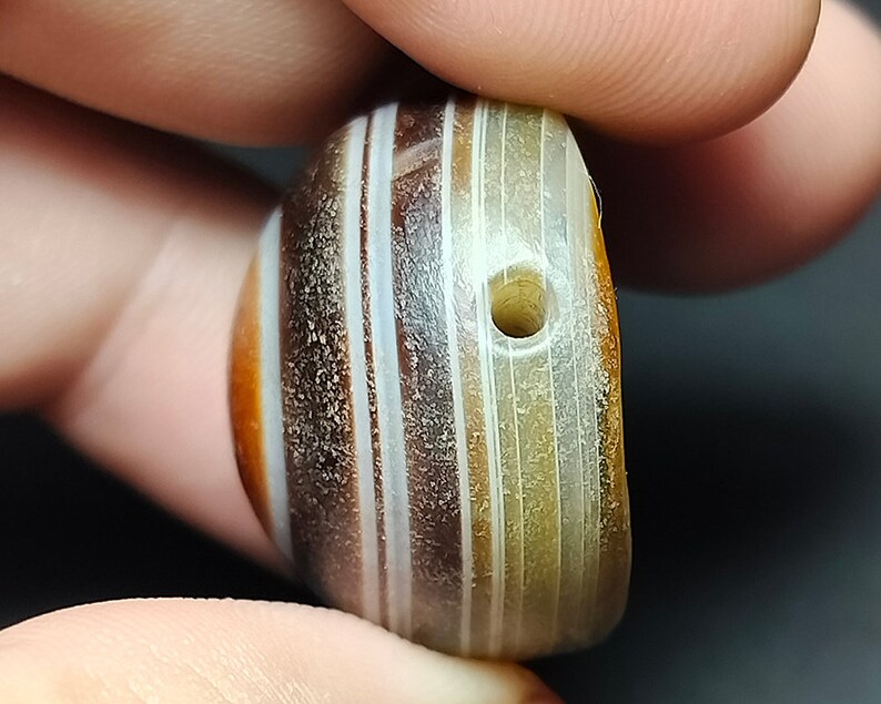 May include: A round, striped agate bead with a hole drilled through the center. The bead is a mix of brown, white, and cream colors.