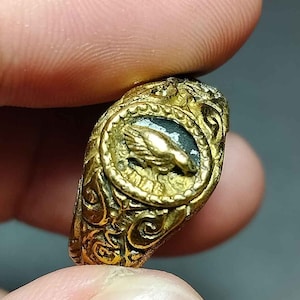 Può includere: Un anello d'oro con un designo intricato che presenta un uccello in una cornice circolare. L'anello ha un motivo dettagliato e ornato.