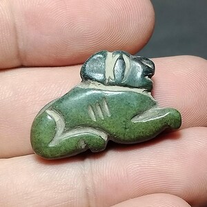 Puede incluir: Pequeña figura de piedra tallada verde de un animal sentado, posiblemente un perro o un león. La figura tiene un diseño detallado con líneas talladas y un acento de color claro en la cabeza. La piedra tiene un aspecto moteado.