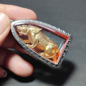 Peut inclure: Pendentif doré représentant un lion stylisé et détaillé, enfermé dans un cadre rectangulaire transparent. Le cadre est bordé d'un métal argenté au design orné. Le pendentif est tenu par une main sur fond sombre.