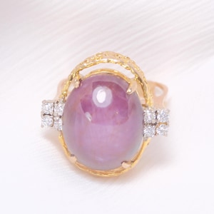 May include: A gold ring featuring a large, oval, purplish-pink gemstone. The ring has a decorative gold setting with small, square clusters of white gemstones on each side of the main stone. The ring is set against a white background.
