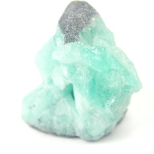 May include: A close-up of a light green mineral specimen with a dark gray top. The mineral has a crystalline structure and a rough texture. The background is white.