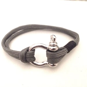 May include: A gray paracord bracelet with a silver metal clasp. The clasp is a large, round ring with a small loop on top.