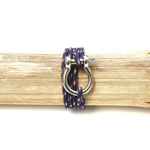 May include: A navy blue and red rope bracelet with a silver metal clasp. The bracelet is wrapped around a piece of wood.