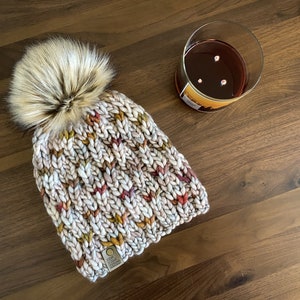May include: A knitted beanie with a fluffy pom pom in shades of beige, brown, and red. The beanie has a small brown tag with the text "The Yarn Whisperer" on it.