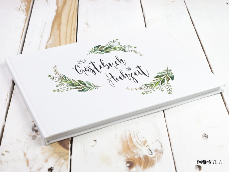 May include: White guest book with a green leafy wreath and the text "Unser G&auml;stebuch zur Hochzeit" in black cursive.