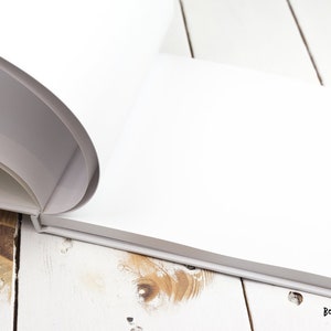May include: A white blank book with a rounded spine is open and lying on a white wooden surface. The book is closed on the right side and the pages are visible on the left side. The book is lying on a white wooden surface with a light brown wood grain.