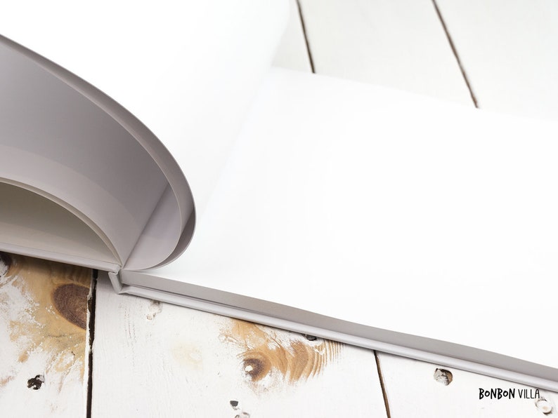 May include: A white blank book with a rounded spine is open and lying on a white wooden surface. The book is closed on the right side and the pages are visible on the left side. The book is lying on a white wooden surface with a light brown wood grain.