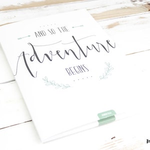 Könnte beinhalten: Weiße Karte mit dem Text "And so the adventure begins" in schwarzer Kursivschrift. Die Karte hat ein grünes Blattdesign und einen kleinen grünen Aufkleber.