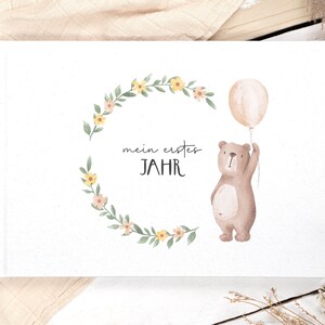 May include: A white photo album with a floral wreath and a brown bear holding a balloon. The text "mein erstes JAHR" is written on the album.