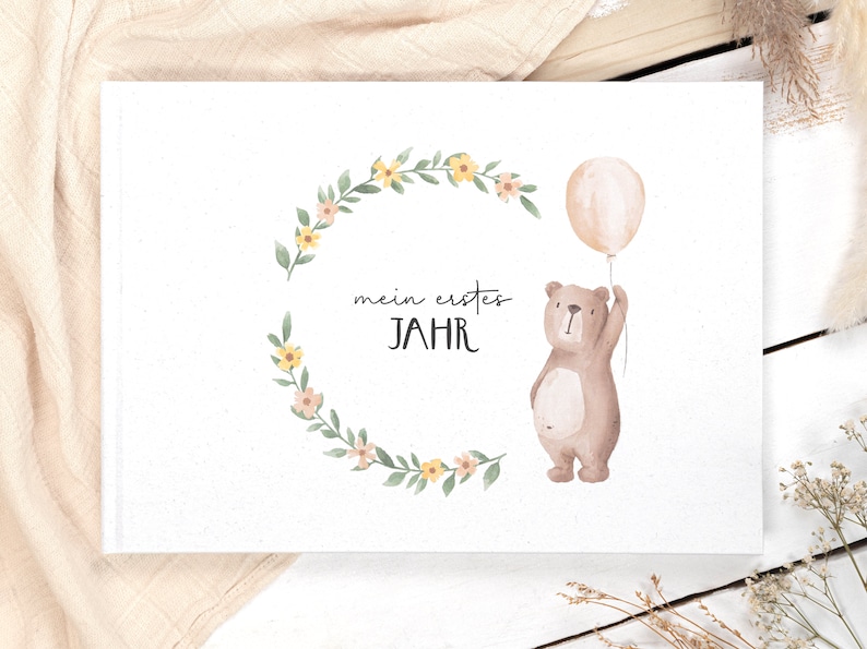 May include: A white photo album with a floral wreath and a brown bear holding a balloon. The text "mein erstes JAHR" is written on the album.