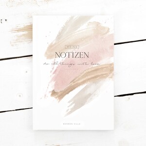 Könnte beinhalten: Ein weißes Notizbuch mit einem rosa und braunen Aquarelldesign. Der Text "MEINE NOTIZEN do all things with love" ist auf dem vorderen Umschlag gedruckt. Das Notizbuch liegt auf einer weißen Holzoberfläche.