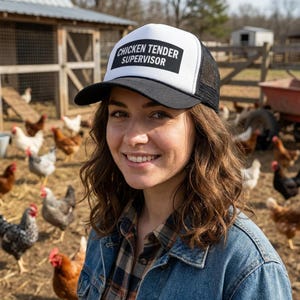 May include: A black and white trucker hat with the text "CHICKEN TENDER SUPERVISOR" in a black rectangle. The hat is worn by a person in a denim jacket, with chickens in the background.