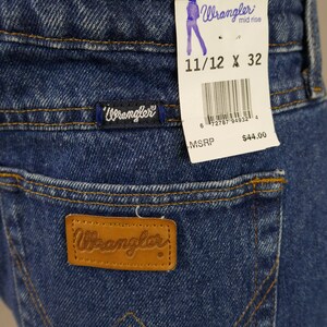 May include: Close-up of blue denim Wrangler Cowboy Cut jeans. The jeans have a brown leather patch with the Wrangler logo on the back pocket. A tag indicates size 11/12 x 32.