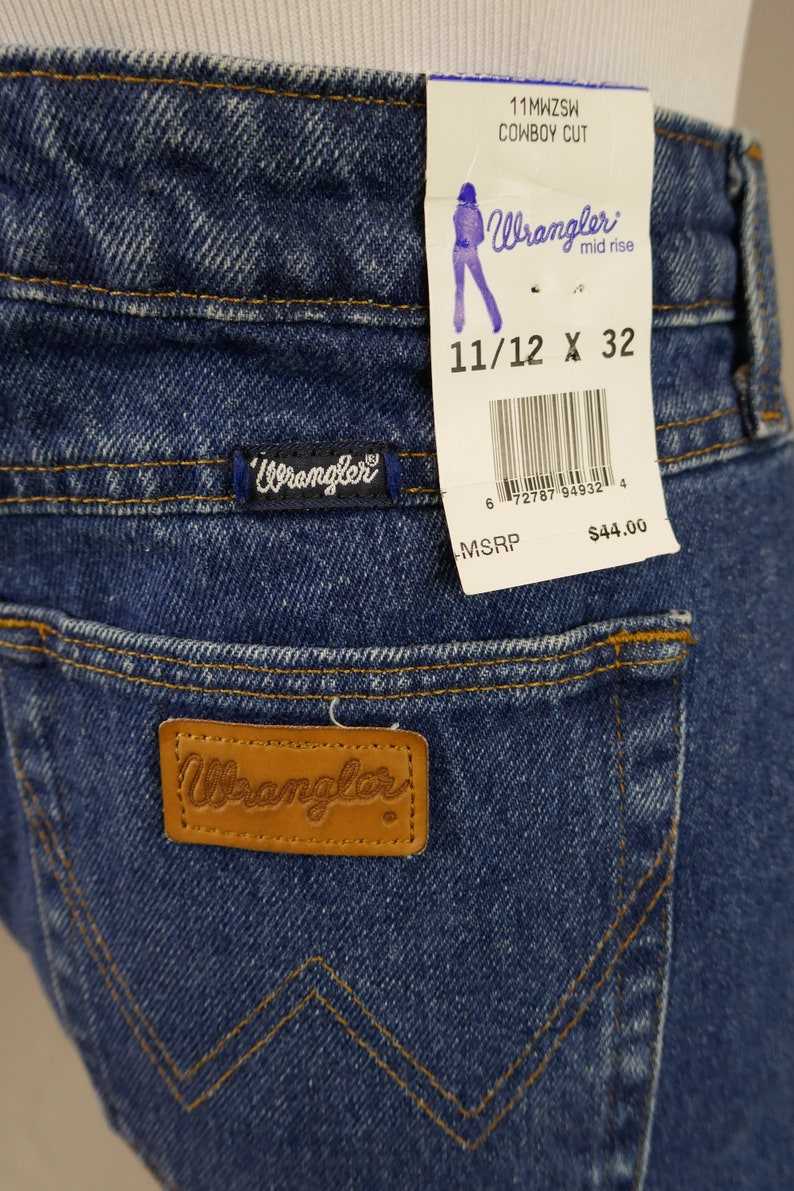 May include: Close-up of blue denim Wrangler Cowboy Cut jeans. The jeans have a brown leather patch with the Wrangler logo on the back pocket. A tag indicates size 11/12 x 32.