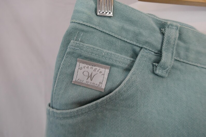 K&ouml;nnte beinhalten: Nahaufnahme einer hellt&uuml;rkisen Jeans. Die Jeans hat eine Vordertasche mit einem kleinen Etikett mit der Aufschrift "Wrangler for women". Die Jeans h&auml;ngt an einem Holzkleiderb&uuml;gel.