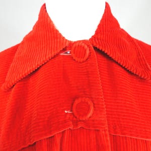 May include: A bright red corduroy jacket featuring a wide pointed collar and two large, fabric-covered circular buttons down the front. The garment is constructed with a distinct vertical ribbed texture throughout the entire surface of the fabric.