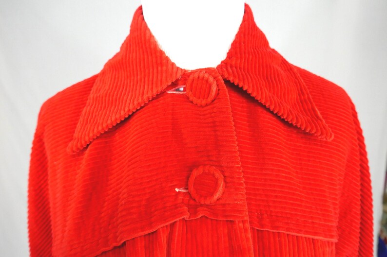 May include: A bright red corduroy jacket featuring a wide pointed collar and two large, fabric-covered circular buttons down the front. The garment is constructed with a distinct vertical ribbed texture throughout the entire surface of the fabric.