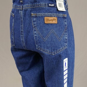 May include: Blue denim Wrangler jeans with a classic five-pocket design. Features a brown leather patch with the Wrangler logo and a tag indicating size 11/12 x 32. The jeans have a straight leg cut.