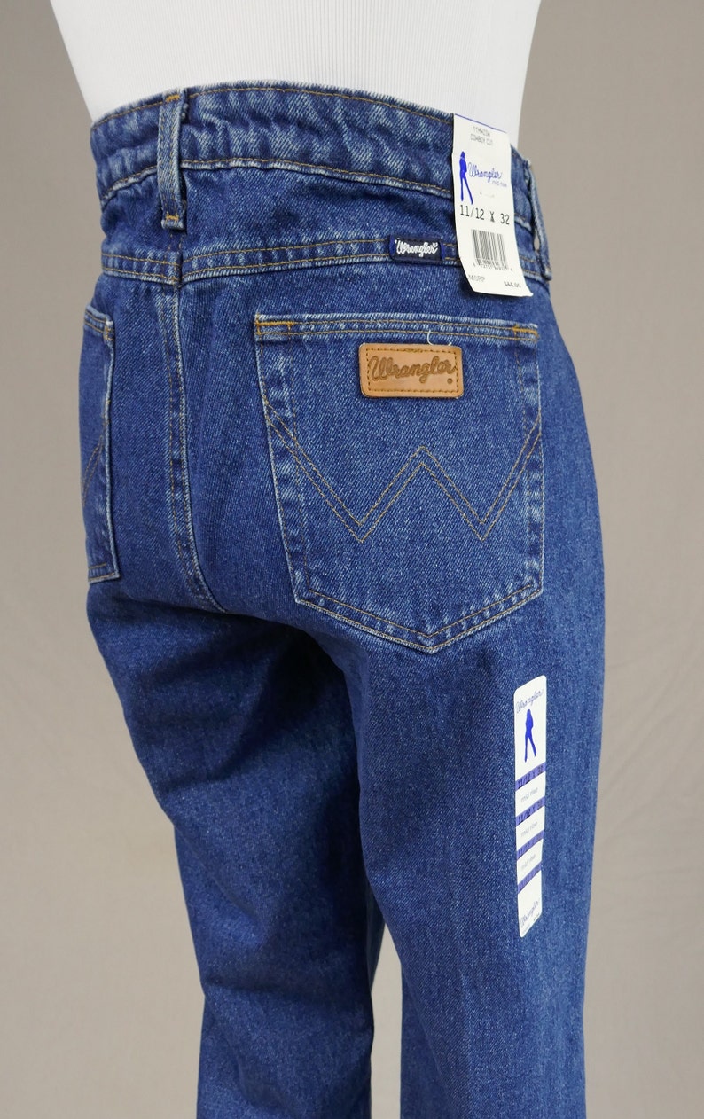May include: Blue denim Wrangler jeans with a classic five-pocket design. Features a brown leather patch with the Wrangler logo and a tag indicating size 11/12 x 32. The jeans have a straight leg cut.