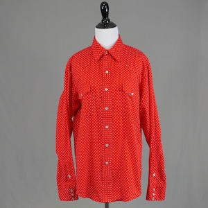 May include: A bright red button-down shirt with a white polka dot pattern. The long-sleeved shirt has two chest pockets and buttoned cuffs. The shirt is displayed on a mannequin against a gray background.