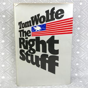 以下が含まれることがあります： トム・ウルフ著『The Right Stuff』のハードカバー本。白いダストジャケットには大きな黒い文字が印刷されており、白い鷲が描かれた青い長方形と、赤と白の水平なストライプからなる旗のようなグラフィックがデザインされています。