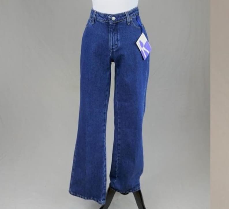 May include: Dark blue denim jeans with a high waist and flared leg. The jeans have a button and zipper closure, belt loops, and pockets. A tag is attached to the jeans. The jeans are displayed on a mannequin.