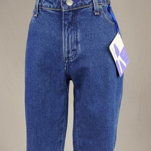 May include: Close-up of blue denim jeans with a button closure and a small tag attached to the front pocket. The jeans are displayed on a mannequin, showcasing the high-waisted design and straight leg fit.