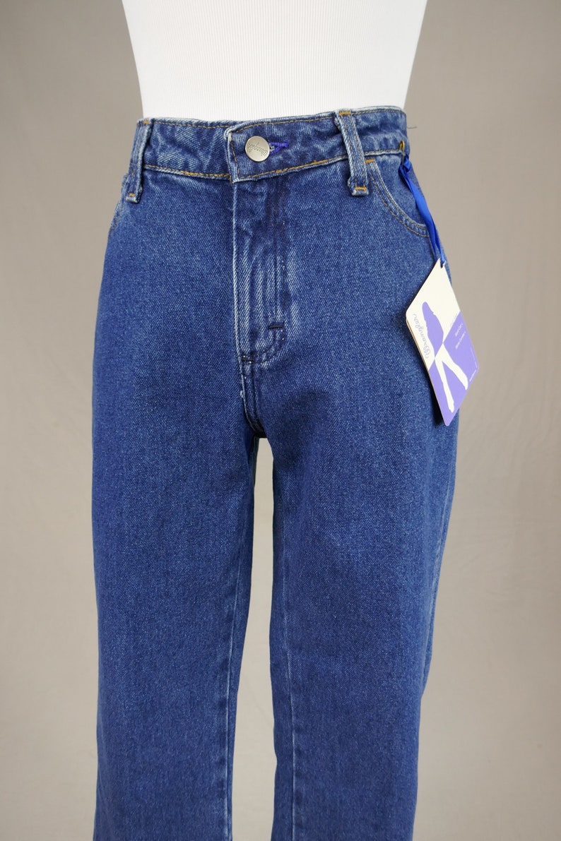 May include: Close-up of blue denim jeans with a button closure and a small tag attached to the front pocket. The jeans are displayed on a mannequin, showcasing the high-waisted design and straight leg fit.
