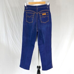 Peut inclure: Jean en denim bleu foncé à taille haute avec des surpiqûres orange contrastantes, deux poches arrière à rabats pointus, un écusson en similicuir beige sur la poche droite, des passants de ceinture et une coupe droite sur cintre en bois.