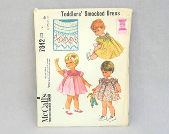 May include: A McCall's 7842 sewing pattern envelope for a toddlers' smocked dress, size 2. The front features illustrations of three children wearing different smocked dresses in yellow, pink, and floral patterns, with a detail of the smocking.