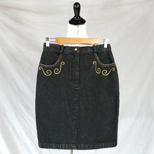 May include: A black denim skirt featuring a high-waisted design with belt loops and a front button closure. The two front pockets are embellished with silver-toned studs and yellow embroidered swirl patterns along the pocket edges.