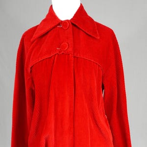 May include: A bright red corduroy jacket featuring a wide collar, long sleeves, and a front button closure with two matching fabric-covered buttons. The garment has a horizontal seam detail across the chest and a textured, vertical ribbed fabric.