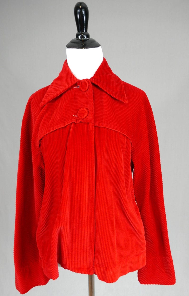 May include: A bright red corduroy jacket featuring a wide collar, long sleeves, and a front button closure with two matching fabric-covered buttons. The garment has a horizontal seam detail across the chest and a textured, vertical ribbed fabric.