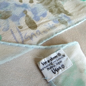 May include: A sheer scarf with a light blue, green, and purple floral pattern. A white fabric tag is attached, reading: Vera-sheer, 62% vinal, 38% silk, Made in Japan, Vera. The scarf features a light blue rolled hem and a signature printed on the fabric.