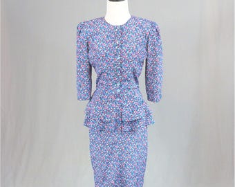 May include: A two-piece set consisting of a long-sleeved blouse and a knee-length skirt, both featuring a dense floral pattern in shades of blue, pink, and white. The blouse has a round neckline, buttons down the front, and a double-tiered peplum waist.