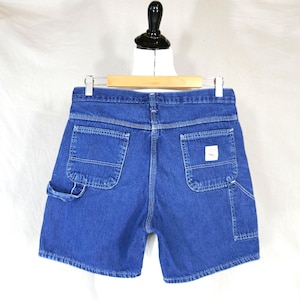 May include: Blue denim shorts with multiple pockets, including a small square pocket on the back. The shorts are hanging on a wooden hanger against a white background. The shorts have white stitching.