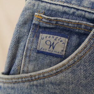 May include: Close-up of a light blue denim jean pocket. The pocket features a blue and white label with the "Wrangler" logo and the text "for WOMEN". The pocket is stitched with yellow thread.