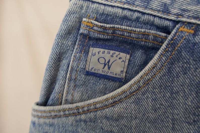 May include: Close-up of a light blue denim jean pocket. The pocket features a blue and white label with the "Wrangler" logo and the text "for WOMEN". The pocket is stitched with yellow thread.