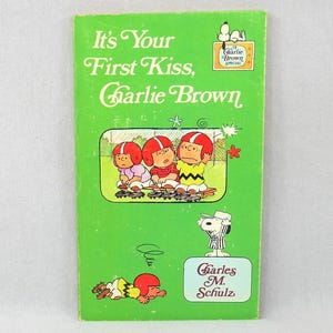 May include: A vintage paperback book titled "It's Your First Kiss, Charlie Brown" with a green cover. The cover features illustrations of Charlie Brown and friends in football helmets. The book also includes the text "A Charlie Brown Special."