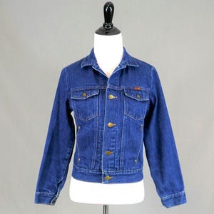 May include: A dark blue denim jacket with a classic design. The jacket features two chest pockets, two side pockets, and a button-up front. The jacket is displayed on a mannequin. The jacket has a small orange tag on the chest pocket.