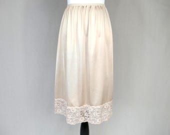 May include: A beige knee-length half slip featuring a smooth, satin-like fabric with a gathered elastic waistband and a wide, intricate floral lace trim along the bottom hem. The garment has a simple, straight silhouette with vertical folds.