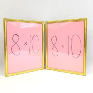 May include: A double hinged picture frame with a metallic gold finish and a textured border. Each side holds an 8x10 inch insert, and the pink paper inserts feature the handwritten text 8x10 in dark ink on both sides of the frame.