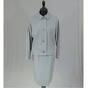 May include: A light gray skirt suit displayed on a mannequin. The suit includes a collared jacket with three buttons and two pockets, paired with a matching knee-length skirt. The buttons are silver-toned.