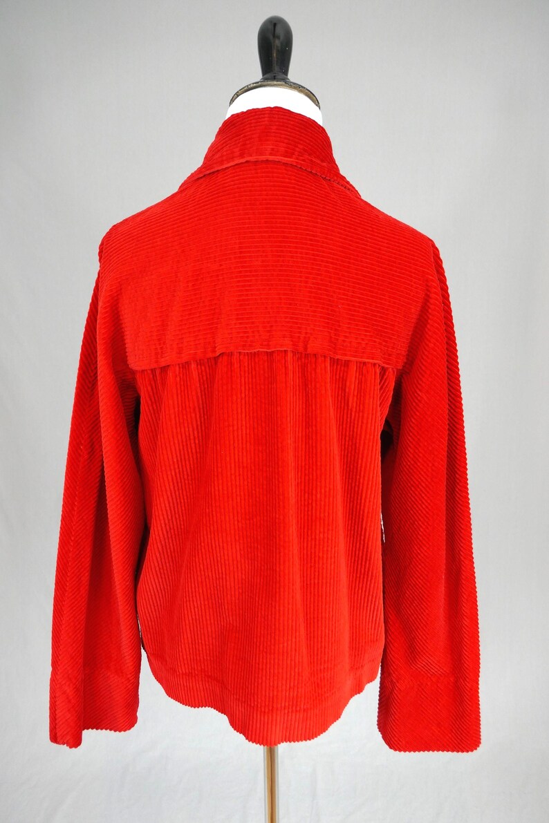 May include: A bright red corduroy jacket featuring a classic collar, long sleeves, and a horizontal back yoke seam. The garment is constructed from a textured, vertically ribbed fabric and is displayed on a mannequin to show its structure.