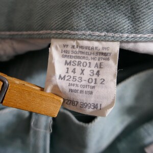 K&ouml;nnte beinhalten: Nahaufnahme einer hellgr&uuml;nen Jeans mit einem wei&szlig;en Etikett. Das Etikett lautet "VF JEANSWEAR, INC" und enth&auml;lt die Gr&ouml;&szlig;e "14 X 34". Die Jeans besteht aus 100 % Baumwolle und wurde in den USA hergestellt.