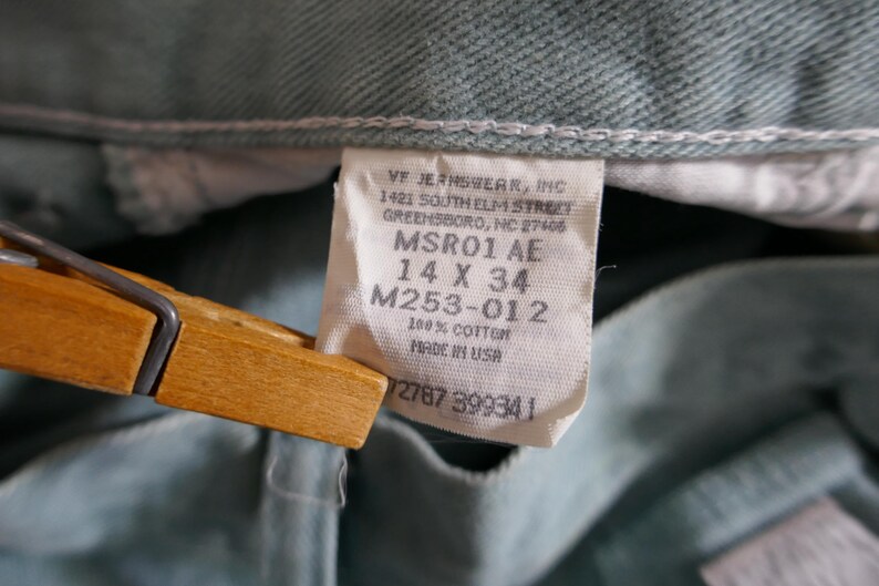 K&ouml;nnte beinhalten: Nahaufnahme einer hellgr&uuml;nen Jeans mit einem wei&szlig;en Etikett. Das Etikett lautet "VF JEANSWEAR, INC" und enth&auml;lt die Gr&ouml;&szlig;e "14 X 34". Die Jeans besteht aus 100 % Baumwolle und wurde in den USA hergestellt.