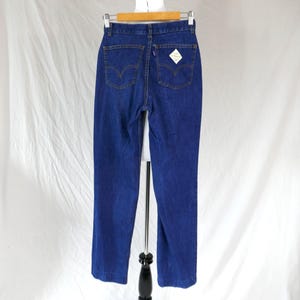 May include: A pair of dark blue denim jeans featuring a high-waisted design, a zipper fly, and two back pockets with a signature arcuate stitch pattern. A small white diamond-shaped tag is attached to the right back pocket, and belt loops.